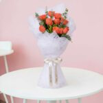 A bouquet of vibrant mix of orange roses and fresh eucalyptus branches, expertly wrapped in a white non-woven paper and finished with a satin ribbon.