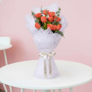 A bouquet of vibrant mix of orange roses and fresh eucalyptus branches, expertly wrapped in a white non-woven paper and finished with a satin ribbon.