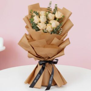 A vibrant bouquet of peach roses is complemented by fresh eucalyptus.The arrangement is wrapped in rustic brown non-woven paper and finished with a sleek black satin ribbon.