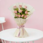 Soft pink roses with fresh green leaves are elegantly wrapped in off-white paper and tied with a vibrant pink ribbon.