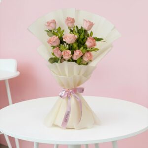 Soft pink roses with fresh green leaves are elegantly wrapped in off-white paper and tied with a vibrant pink ribbon.