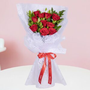 A bouquet of vibrant red roses and fresh green leaves arranged and wrapped in non-woven paper, finished with a bright red satin ribbon.